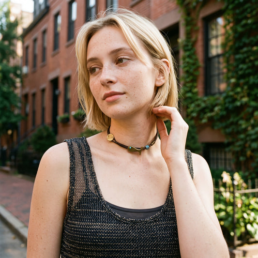 A blonde woman wearing a black beaded choker necklace with gold and blue beads, posing outdoors in front of a brick building with green ivy.
