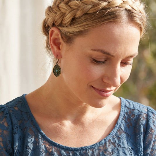 A close-up lifestyle shot of a woman wearing a detailed braided hairpiece and a blue lace top, looking down with a soft smile. She is wearing a dark green oval dangle earring with a gold-toned frame.