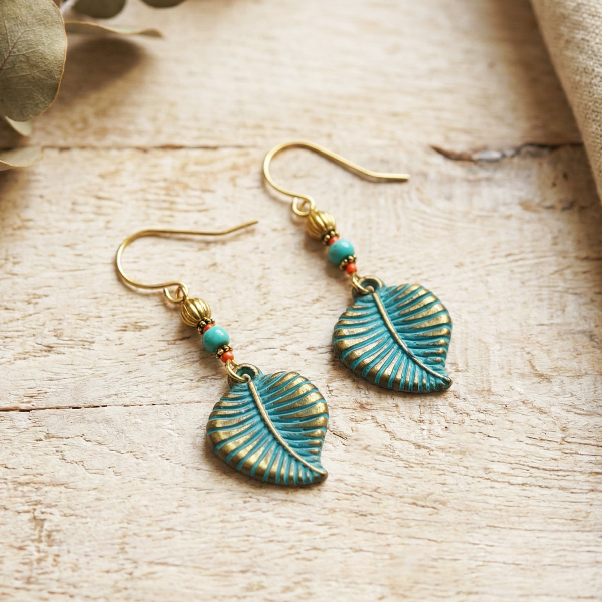 A pair of dangling leaf-shaped earrings with turquoise blue enamel and gold-toned metal, shown on a light wood surface with small turquoise beads and gold hooks.