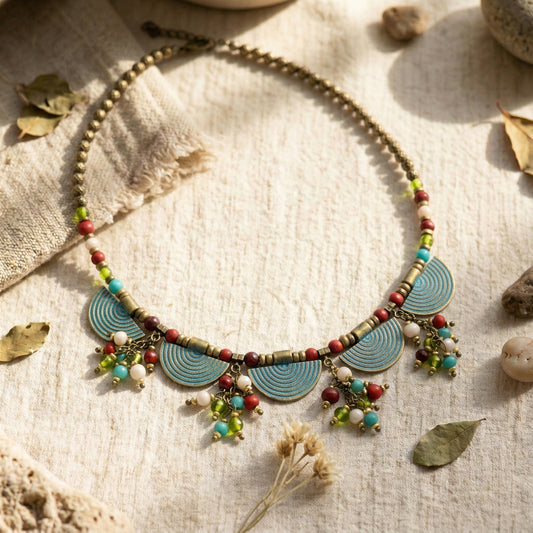 A bohemian-style beaded necklace with turquoise blue spiral discs, bronze-toned beads, and colorful dangling charms, styled on a textured beige linen fabric with dried leaves and small stones.