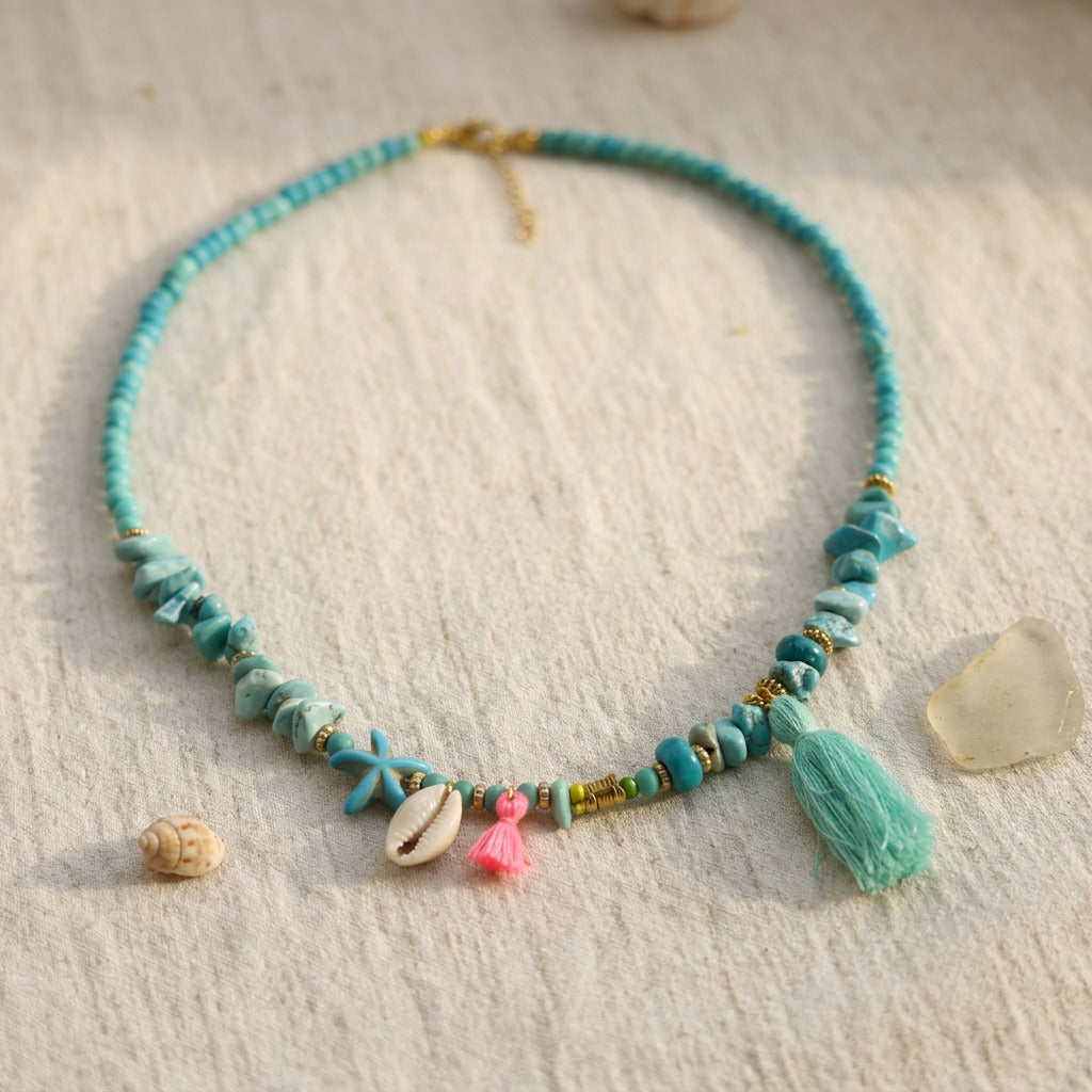 A turquoise beaded necklace with starfish charms, a pink tassel, a cowrie shell, and a large mint green tassel, displayed on a textured cream fabric with seashell and driftwood props.