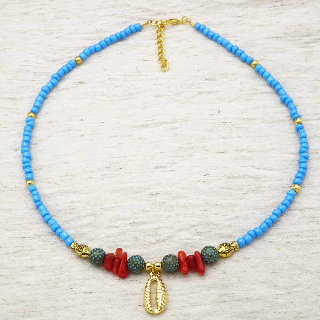 Sky-blue glass seed bead necklace featuring a gold cowrie shell pendant, red coral chips, dotted ceramic beads, and gold fish spacers. Ideal for resort and beach boutiques.