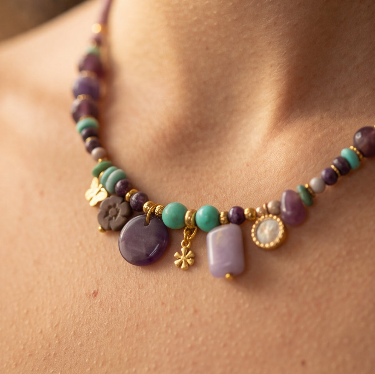 A close-up of a beaded necklace worn on a person's neck, featuring purple and teal beads, gold-toned charms, and a lavender stone pendant.