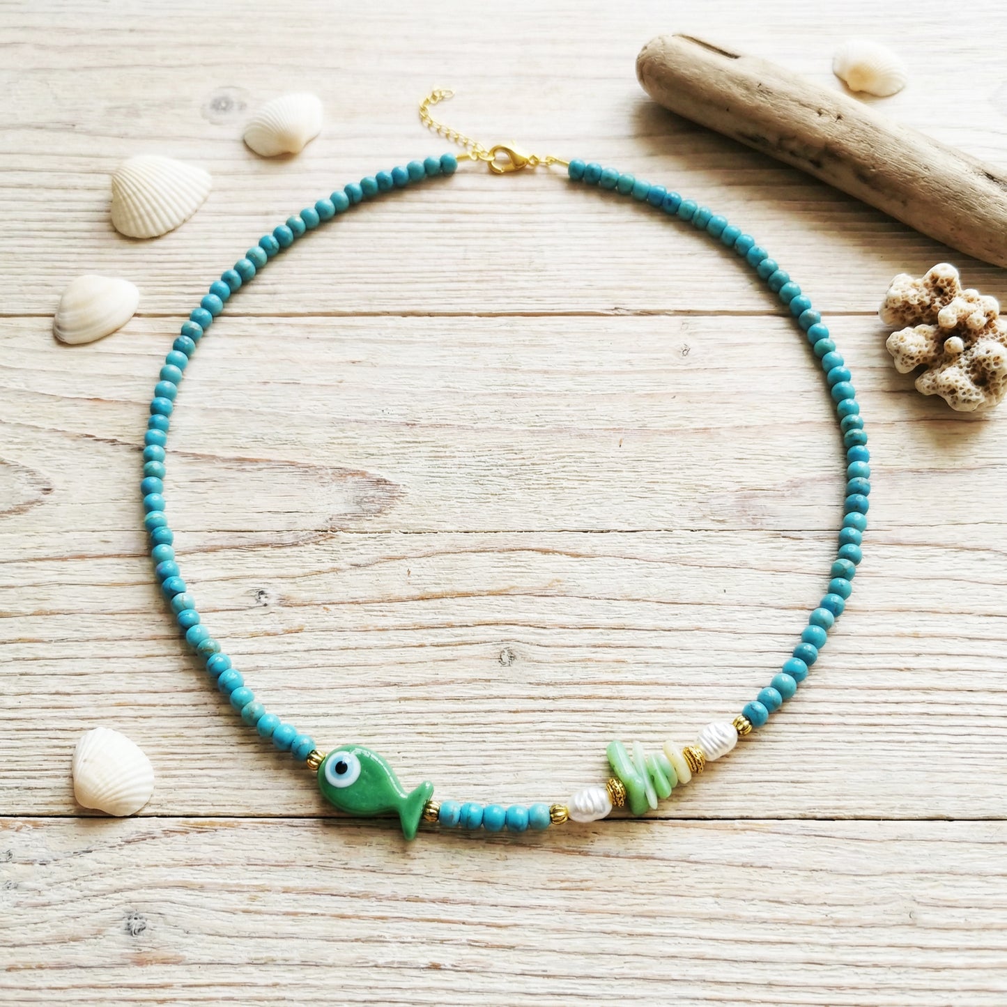 Necklace with blue beads and a green fish charm on a wooden surface with shells and driftwood.