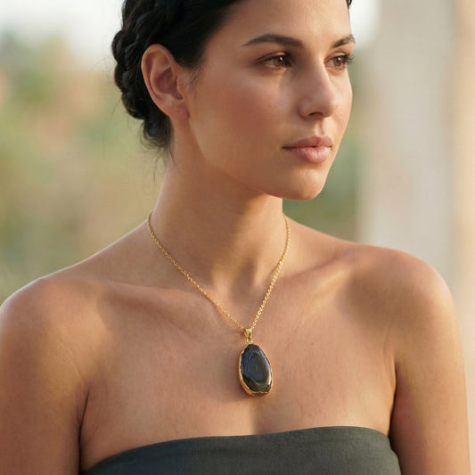 A woman wearing a gold pendant necklace with a dark, polished teardrop-shaped gemstone, shown in a close-up portrait against a softly blurred outdoor background.