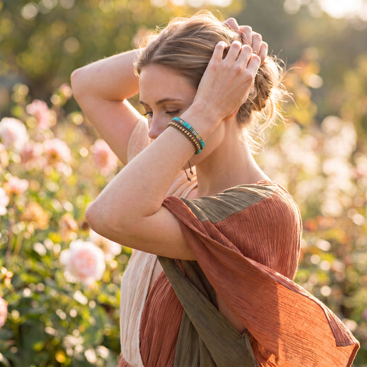 A woman in a rust and olive draped dress poses in a sunlit garden, wearing a stack of beaded bracelets on her wrist.