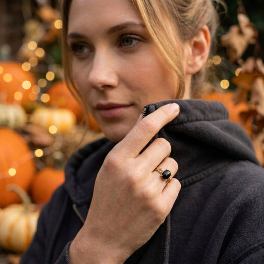 A woman wearing a black onyx ring with a gold band and a dark oval stone, posing in a cozy outdoor setting with pumpkins and warm bokeh lights in the background.