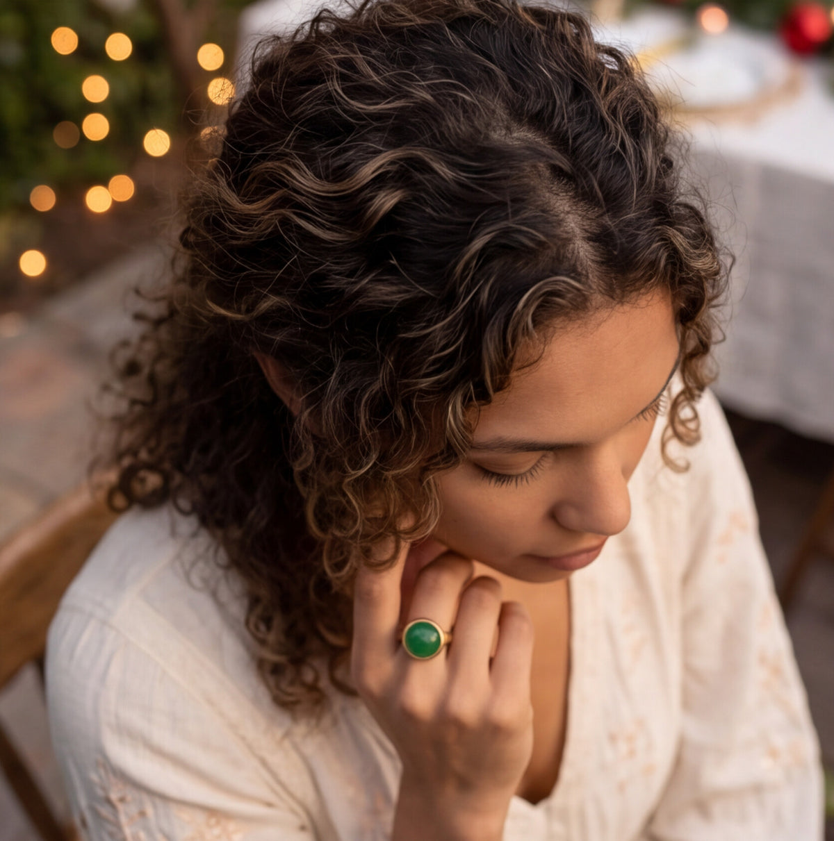 A woman with curly brown hair wearing a white embroidered top and a green ring, captured in a warm, softly lit outdoor setting with blurred lights in the background.