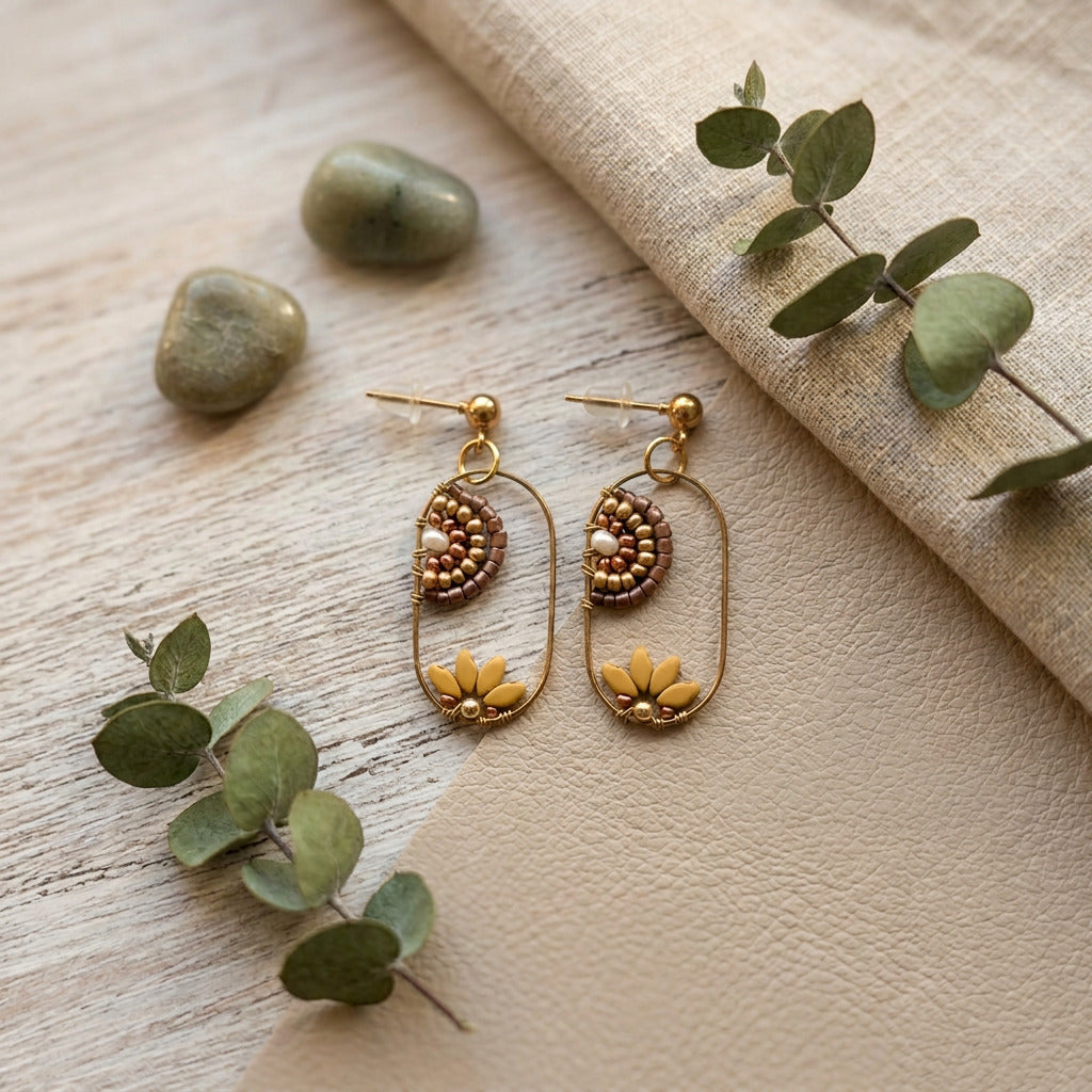 A pair of gold-toned dangle earrings with beaded oval details and small yellow lotus flower charms, styled on a light wood surface with green eucalyptus leaves and a beige fabric runner.