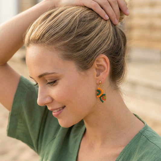 A woman with blonde hair in a bun wearing a pair of handmade beaded earrings shaped like sunflowers, posed in a soft-focus outdoor setting.