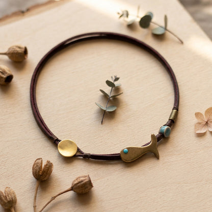 A double-strand dark brown leather necklace with a brass whale-shaped charm, a round brass disc, and turquoise-colored beads, displayed on a light wood background with dried botanicals.