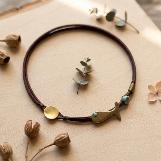 A double-strand dark brown leather necklace with a brass whale-shaped charm, a round brass disc, and turquoise-colored beads, displayed on a light wood background with dried botanicals.