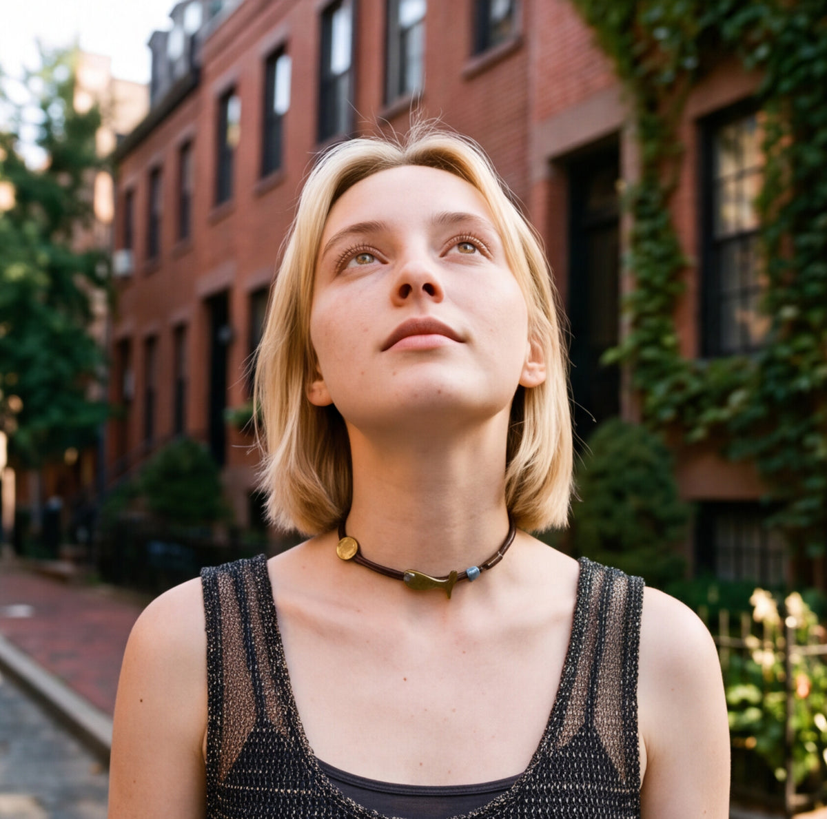 A blonde woman looking upward outdoors, wearing a black mesh tank top and a dark beaded choker necklace with gold and green accents. Brick apartment buildings and greenery are blurred in the background.