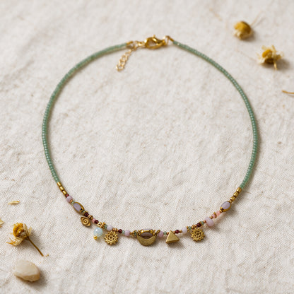 A delicate beaded necklace with a light green cord, gold-toned metal charms, and small pastel beads, displayed on a textured beige fabric with dried flower petals.
