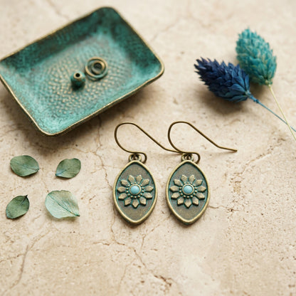 A pair of teardrop-shaped dangle earrings with antique bronze floral medallions and light blue centers, displayed on a textured beige surface with decorative dried flowers and a small green tray.