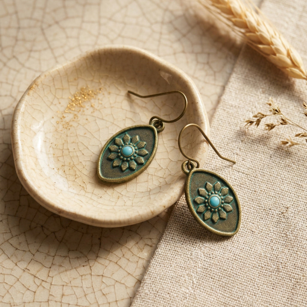 A pair of dangling earrings with oval bronze-toned metal frames, embossed floral details, and a small turquoise center, displayed on a small ceramic dish over a textured beige fabric background.