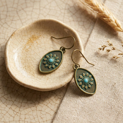 A pair of dangling earrings with oval bronze-toned metal frames, embossed floral details, and a small turquoise center, displayed on a small ceramic dish over a textured beige fabric background.
