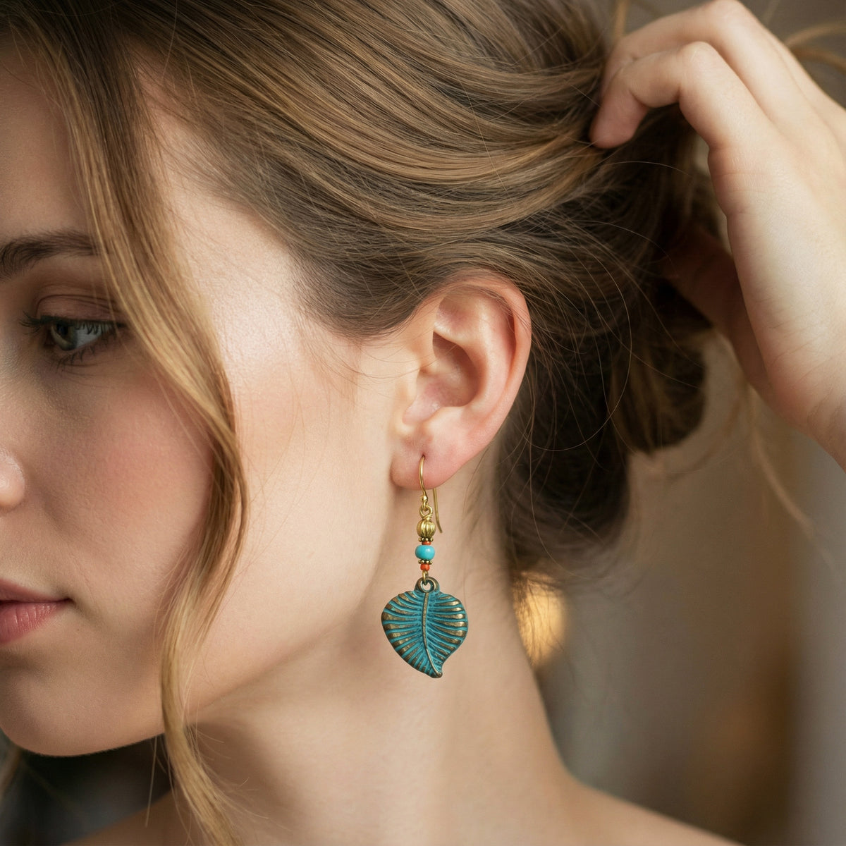 A close-up lifestyle shot of a woman wearing a dangling earring. The earring features a textured turquoise-blue leaf charm with gold-toned metal details and small turquoise beads.