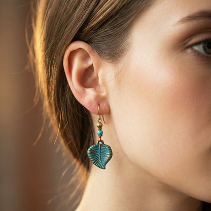 A close-up of a woman's ear wearing a dangling earring with a turquoise-blue leaf charm, gold-toned metal, and small turquoise beads.