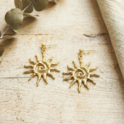 Gold sun-shaped earrings on a wooden surface with leaves and fabric.