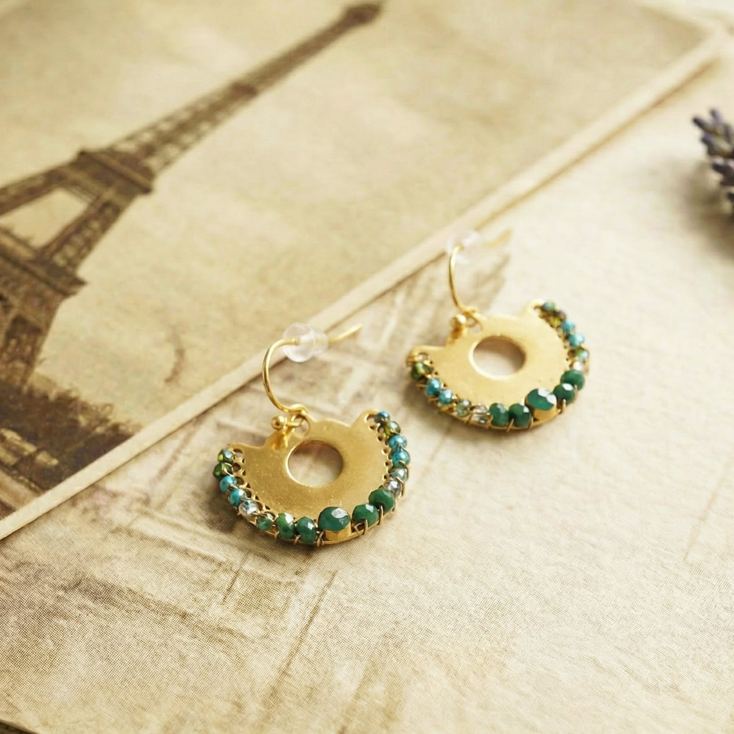 Gold earrings with green gemstones on a textured surface with an Eiffel Tower image.