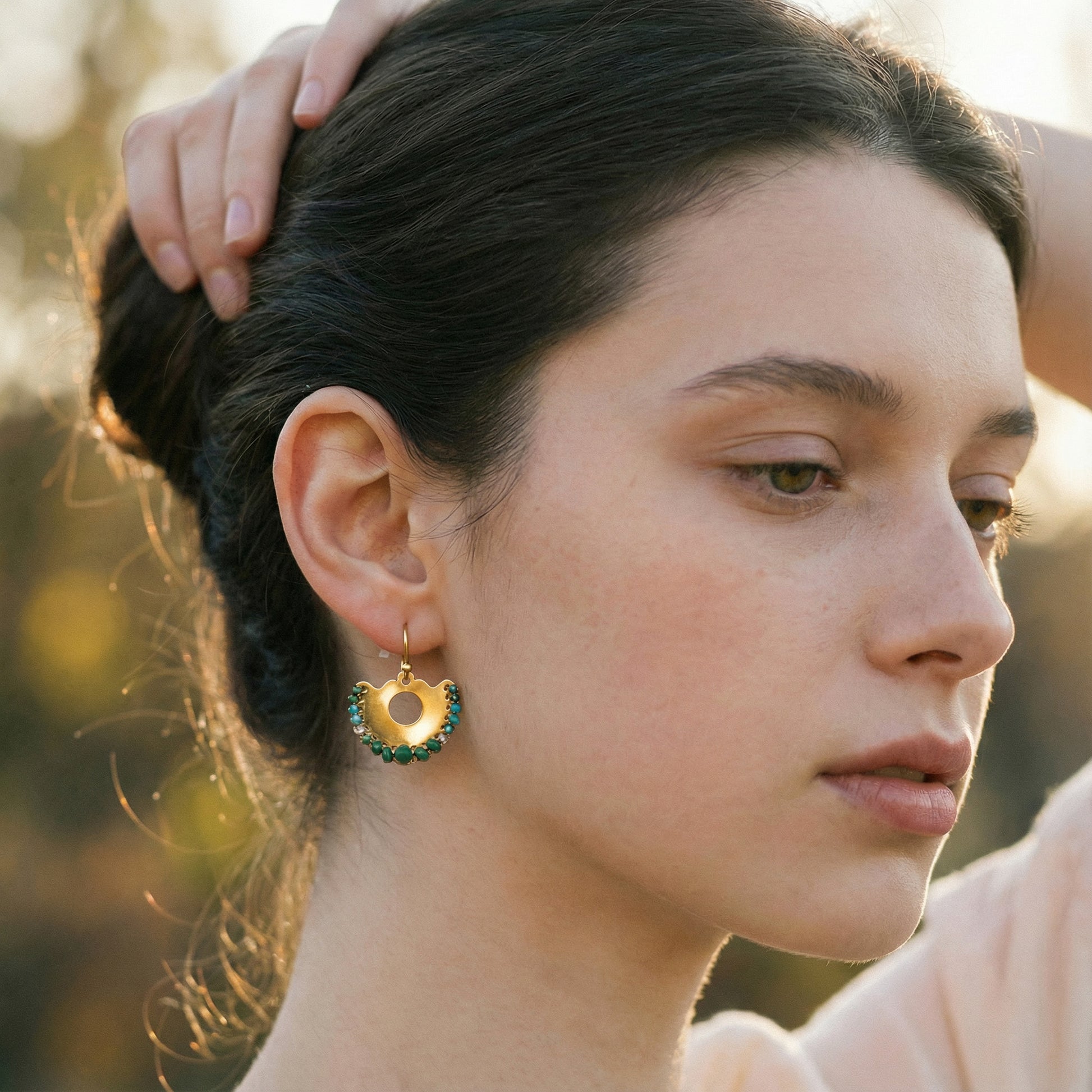 Close-up of a woman wearing gold earrings with green gemstones, outdoors.