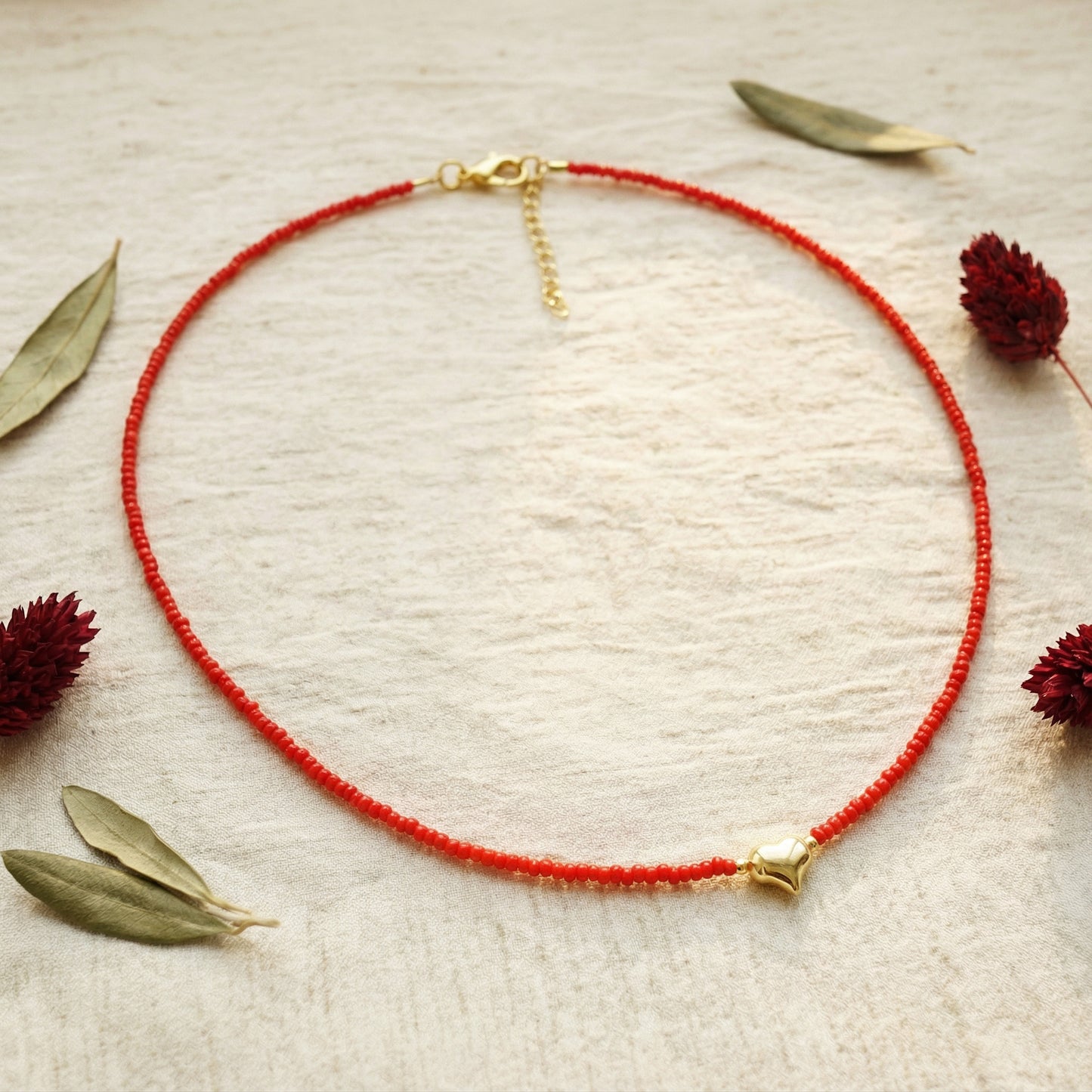 Red beaded necklace with gold clasp on a textured beige surface with dried leaves and flowers.