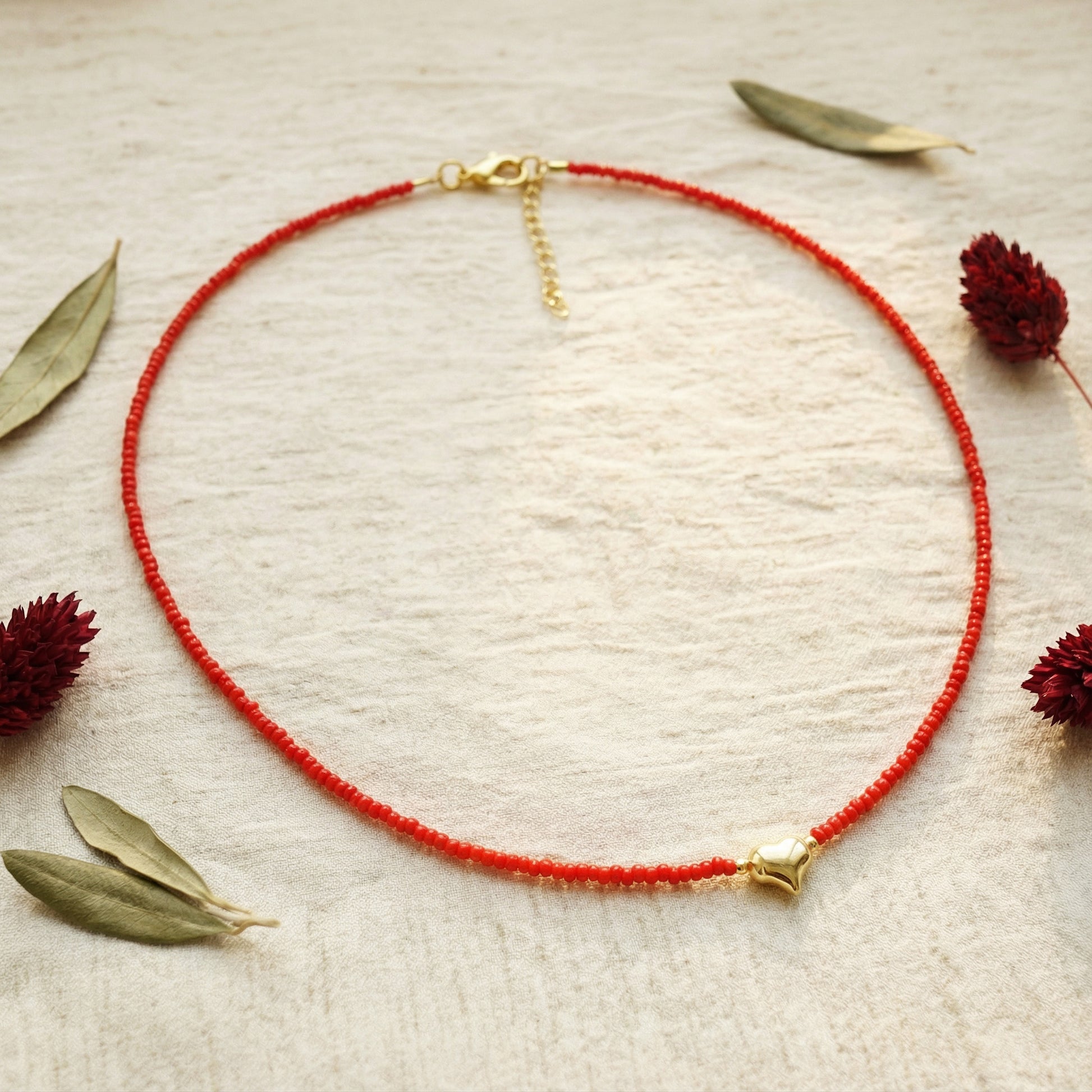 Red beaded necklace with gold clasp on a textured beige surface with dried leaves and flowers.