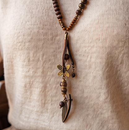 A close-up of a bohemian-style beaded necklace worn on a person's neck, featuring brown wooden beads, a dark brown leather branch pendant with a gold butterfly charm, and a small leaf charm, set against a beige linen shirt.