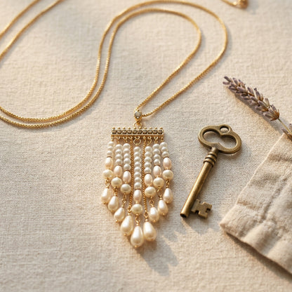A gold-tone necklace with a fringed pearl pendant, styled on a textured beige fabric alongside an antique-style key and dried lavender.