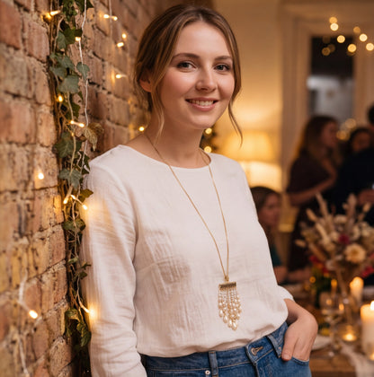 A woman wearing a white long-sleeve top and blue jeans stands against a brick wall decorated with green ivy and warm string lights, wearing a long gold-tone pendant necklace with a pearl-like fringe.