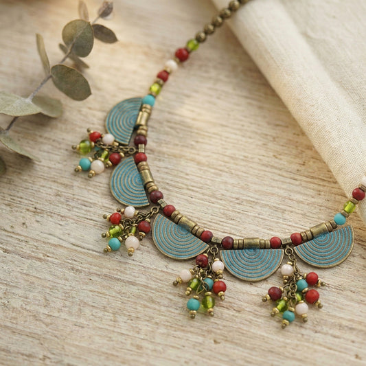 A bohemian-style beaded necklace with turquoise blue semicircle discs, bronze-toned beads, and dangling charms of red, turquoise, green, and white beads, displayed on a light wood surface next to a rolled cream fabric and eucalyptus leaves.