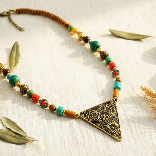 Necklace with colorful beads and a bronze triangle pendant on a textured surface with leaves.