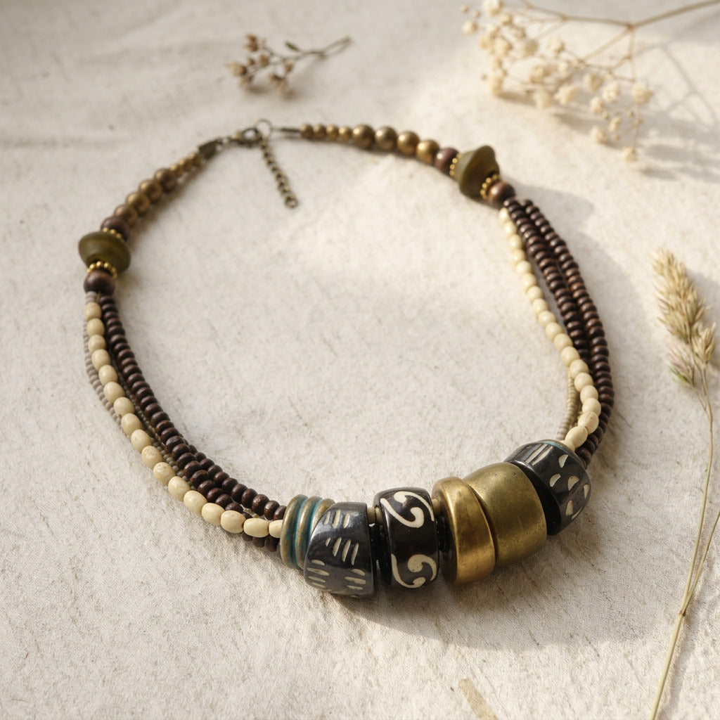 A bohemian-style beaded necklace with dark brown and cream beads, featuring large decorative beads with black, gold, and white patterns, displayed on a textured beige fabric with dried flowers.