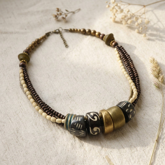 A bohemian-style beaded necklace with dark brown and cream beads, featuring large decorative beads with black, gold, and white patterns, displayed on a textured beige fabric with dried flowers.