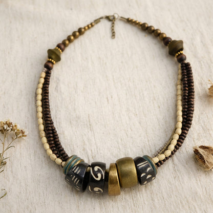 A bohemian-style beaded necklace with dark brown and cream beads, featuring a central cluster of decorative cylindrical beads in black, bronze, and green, displayed on a textured off-white fabric background.