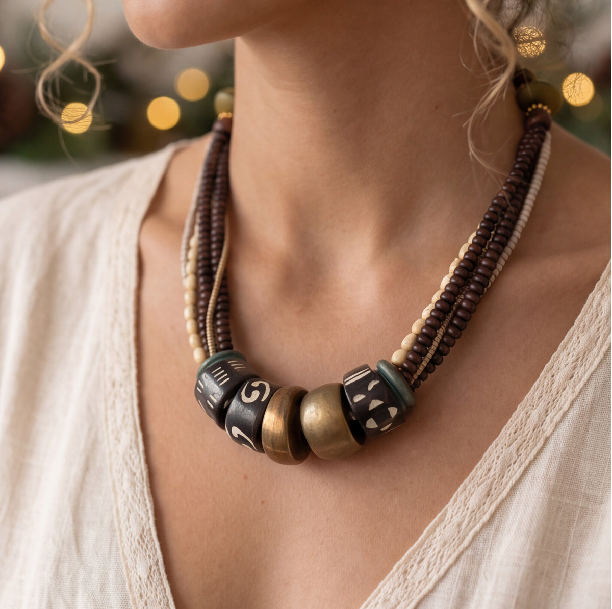 A close-up of a woman wearing a multi-strand statement necklace with dark brown, cream, and bronze beads, featuring decorative cylindrical beads with white lettering.