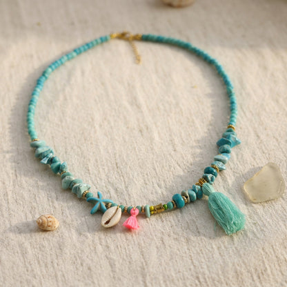 A turquoise beaded necklace with starfish charms, a pink tassel, a cowrie shell, and a large mint green tassel, displayed on a textured cream fabric with seashell and driftwood props.