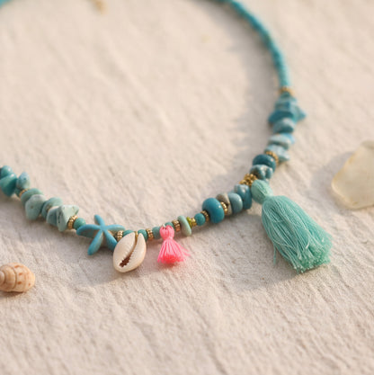 A turquoise and light blue beaded necklace with starfish and shell charms, a pink tassel, and a large mint green tassel, displayed on a textured beige fabric background with small seashell props.