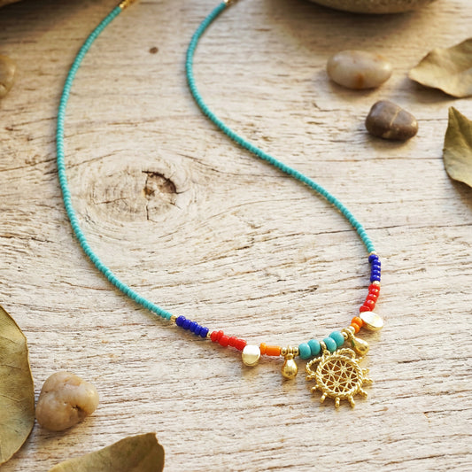 Colorful beaded necklace with a gold pendant on a wooden surface with rocks and leaves.