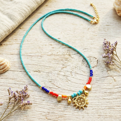 Colorful beaded necklace with a gold pendant on a wooden surface with dried lavender and shells.