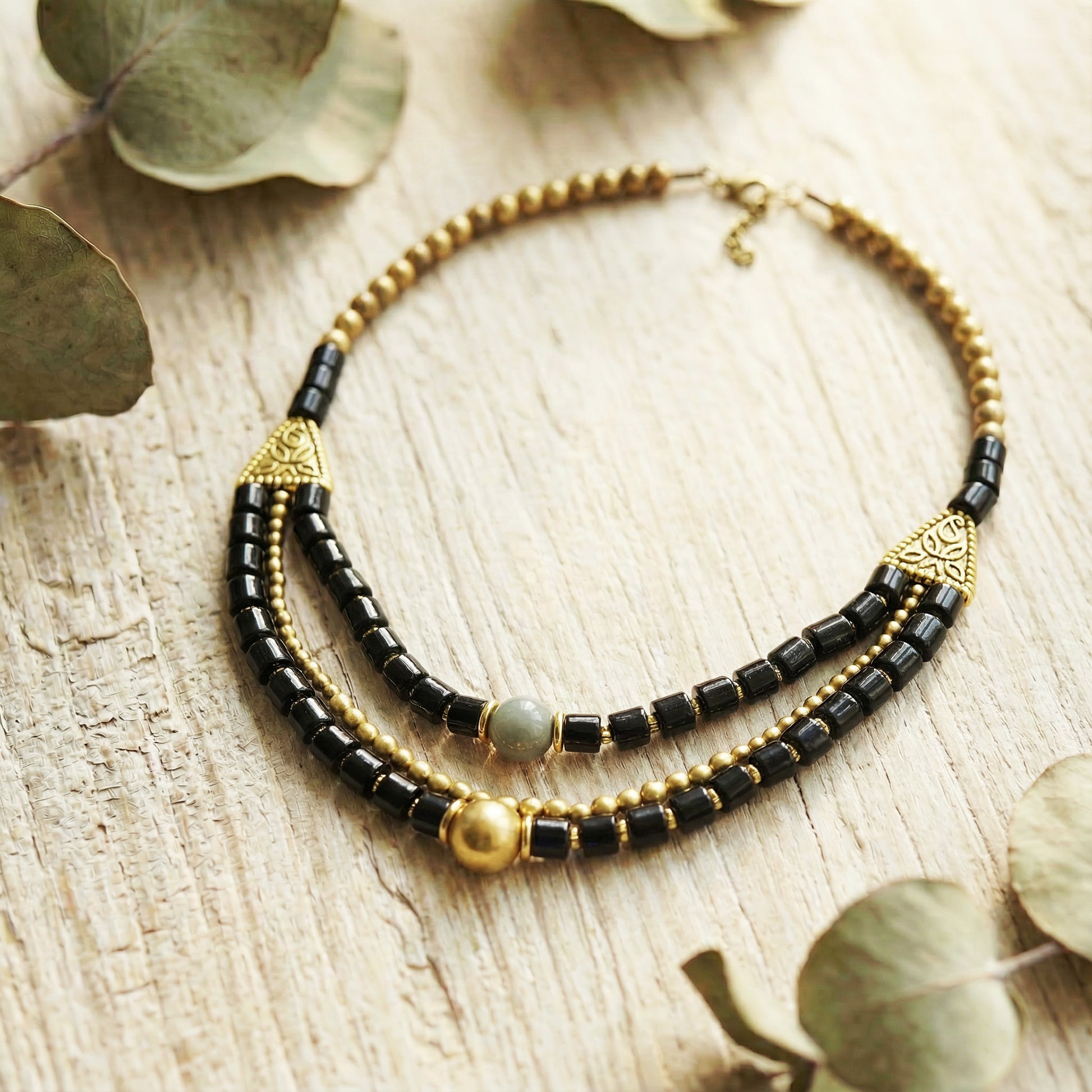 Black and gold beaded necklace on a wooden surface with eucalyptus leaves.