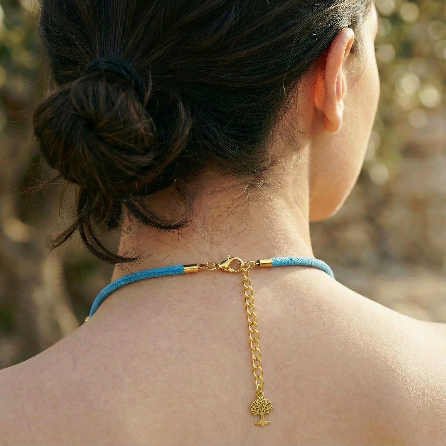 Person wearing a blue choker with a gold chain and tree pendant, outdoors.