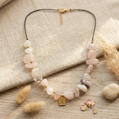 A beaded necklace with light pink and white stone beads, a clear faceted bead, and a small gold pendant, displayed on a beige fabric surface with dried grass and a wood tabletop.