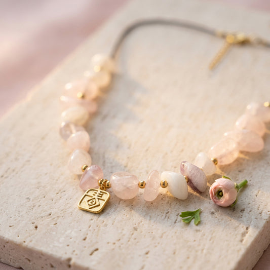 A delicate necklace with light pink and white stone beads, small gold spacer beads, a gold charm, and a small pink flower accent, displayed on a textured beige stone surface.