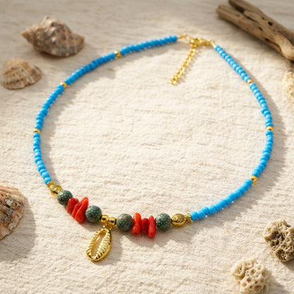 Sky-blue glass seed bead necklace featuring a gold cowrie shell pendant, red coral chips, dotted ceramic beads, and gold fish spacers. Ideal for resort and beach boutiques.