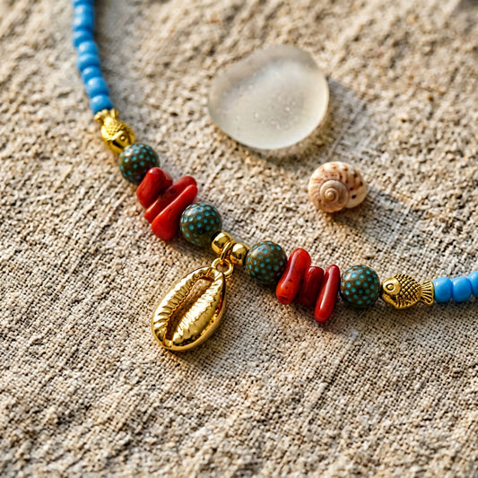 Handcrafted sky blue seed bead coastal necklace featuring a gold cowrie shell charm, coral chips, patterned ceramic beads, and gold fish accents.