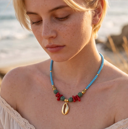 Sky-blue glass seed bead necklace featuring a gold cowrie shell pendant, red coral chips, dotted ceramic beads, and gold fish spacers. Ideal for resort and beach boutiques.