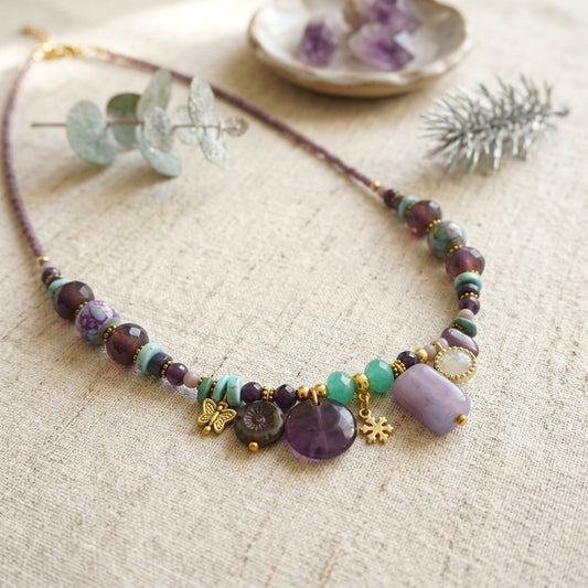 A beaded necklace with purple, green, and lavender beads, gold-toned charms, and a lavender stone pendant, displayed on a textured beige fabric with decorative eucalyptus and amethyst crystals.