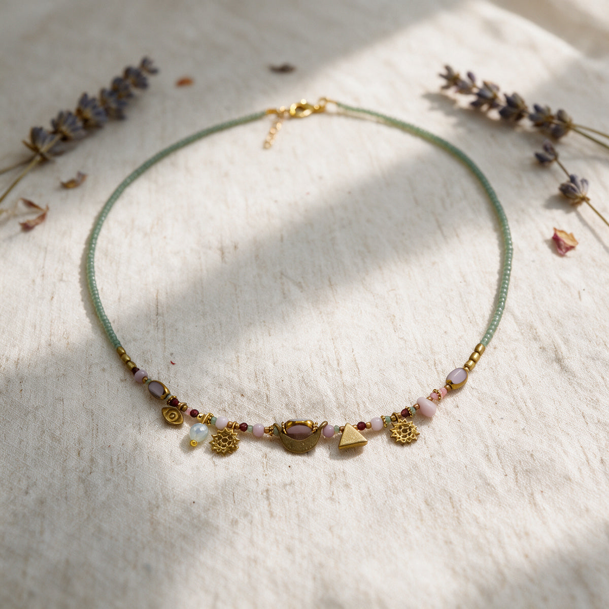 A delicate beaded necklace with a light green cord, gold-toned charms, and small pastel beads, arranged in a circle on a textured off-white surface with dried lavender sprigs in the corners.
